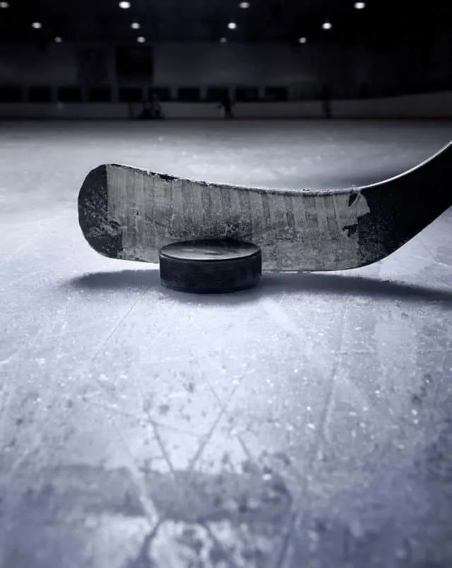 best things to do in Door County in February, Close up shot of a hockey stick resting next to a puck in the scored ice of a hockey stadium with ceiling lights and seating stands visible in the background