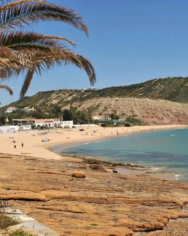day trips from Algarve Portugal, View of curved sandy beach with small collection of buildings next to larger rolling hills covered in green shrubs all under a clear blue sky