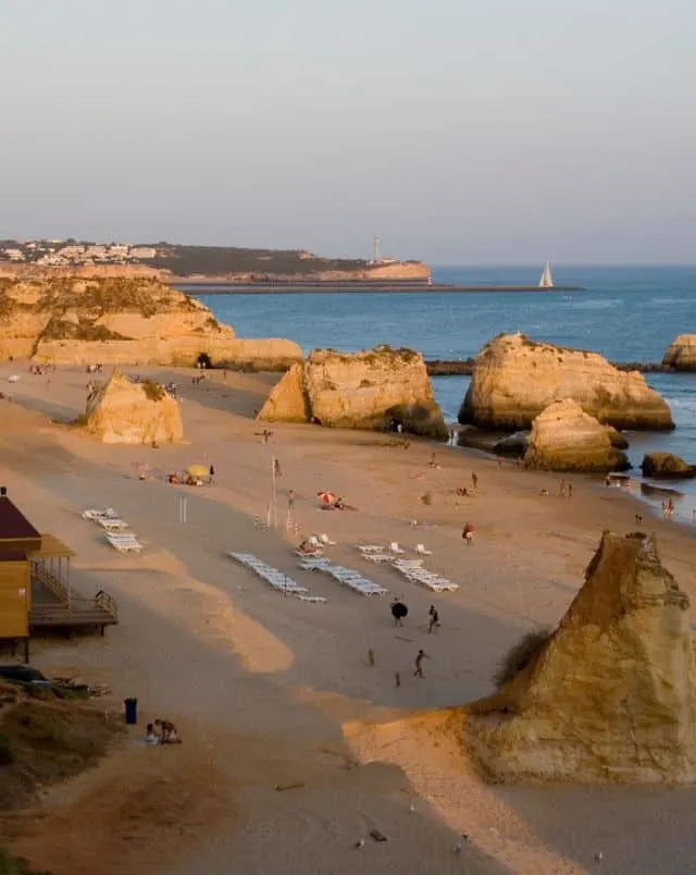 top things to do in Portimao Portugal, View of sandy beach at sunset with people walking amongst beach umbrellas and rows of sun loungers surrounded by tall rock formations sitting on the edge of the ocean with more coastline visible behind
