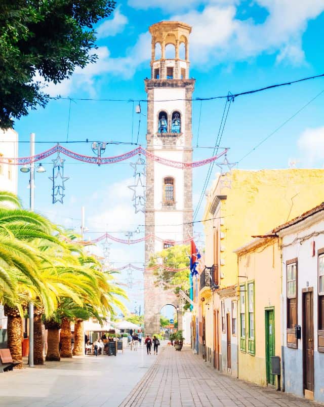 visiting Tenerife in October, View of street with a row of single-level buildings on one side and a row of small green palm trees on the other with a large bell tower visible at the end under a blue sky with clouds