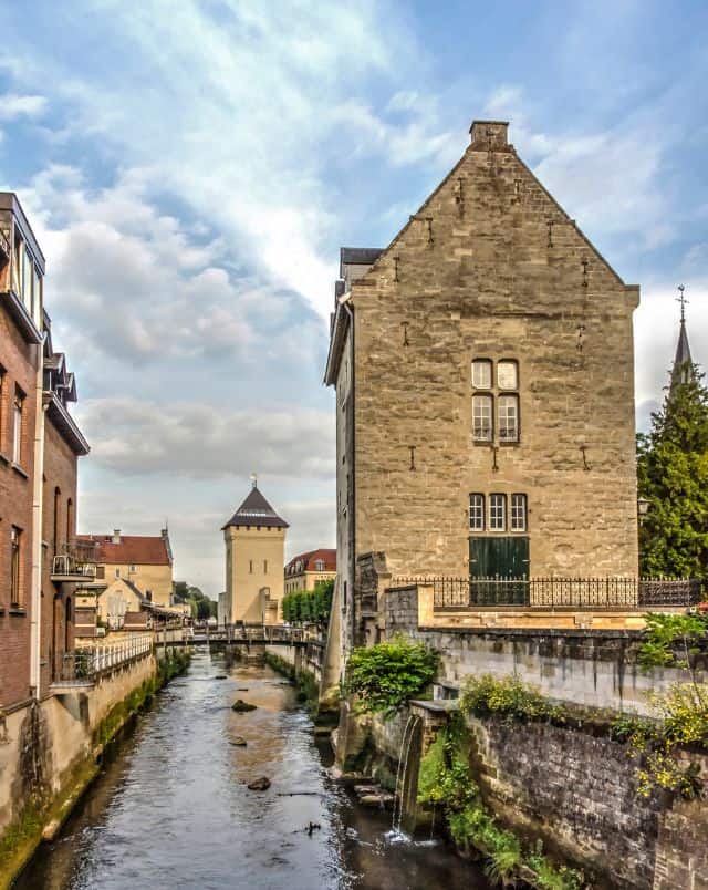 Learn about what to see around Luxembourg for your next trip, view of a small river running through a town with tall stone buildings on either side and a pointed-roofed tower in the distance all under a dramatically cloudy sky