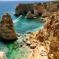 Festive Things to do in Algarve in December, View looking down on a coastline with people sunbathing on a golden sandy beach surrounded by tall rocky cliffs next to clear turquoise seawater
