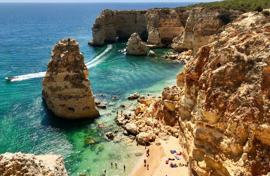 Festive Things to do in Algarve in December, View looking down on a coastline with people sunbathing on a golden sandy beach surrounded by tall rocky cliffs next to clear turquoise seawater