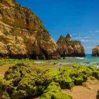 View of rocks covered in virulent green moss standing on sandy beach next to tall rocky cliffs standing on the coast under a bright blue sky
