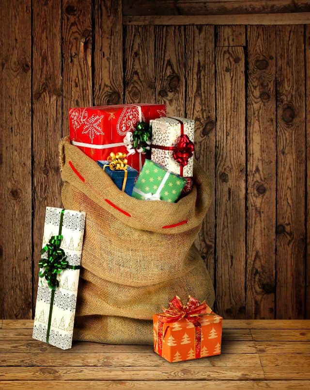 Milwaukee Christmas activities for kids, Close up shot of a hessian bag stuffed full of presents wrapped in colorful paper and ribbons all resting on a wooden bench in front of a wooden wall