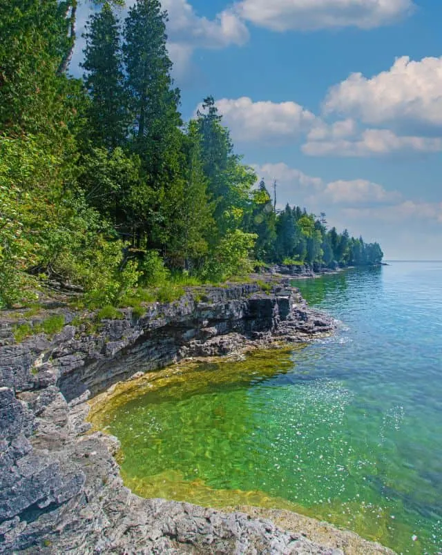 what to do in Door County in March, View of rocky coastline covered in green trees sitting next to a large body of water in Cave Point County Park under a bright blue sky with white fluffy clouds