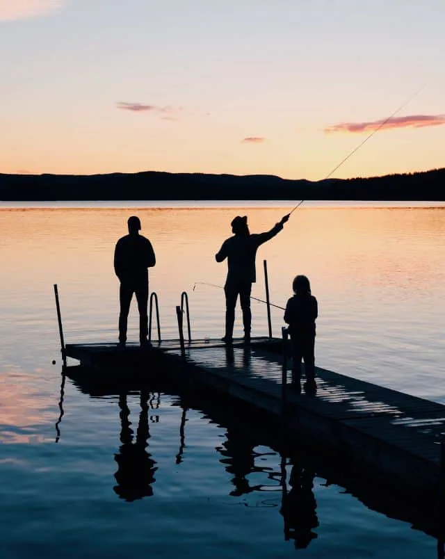 Three people in silhouette standing on a wooden jetty holding fishing rods in front of a calm body of water with some rolling hills visible in the distance at dusk, Northern Wisconsin Fishing Cabins