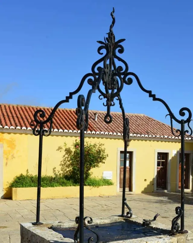 Top Portimao things to do, View of well with metal ornament on top standing in courtyard next to a yellow building with a slate rooftop under a clear azure blue sky