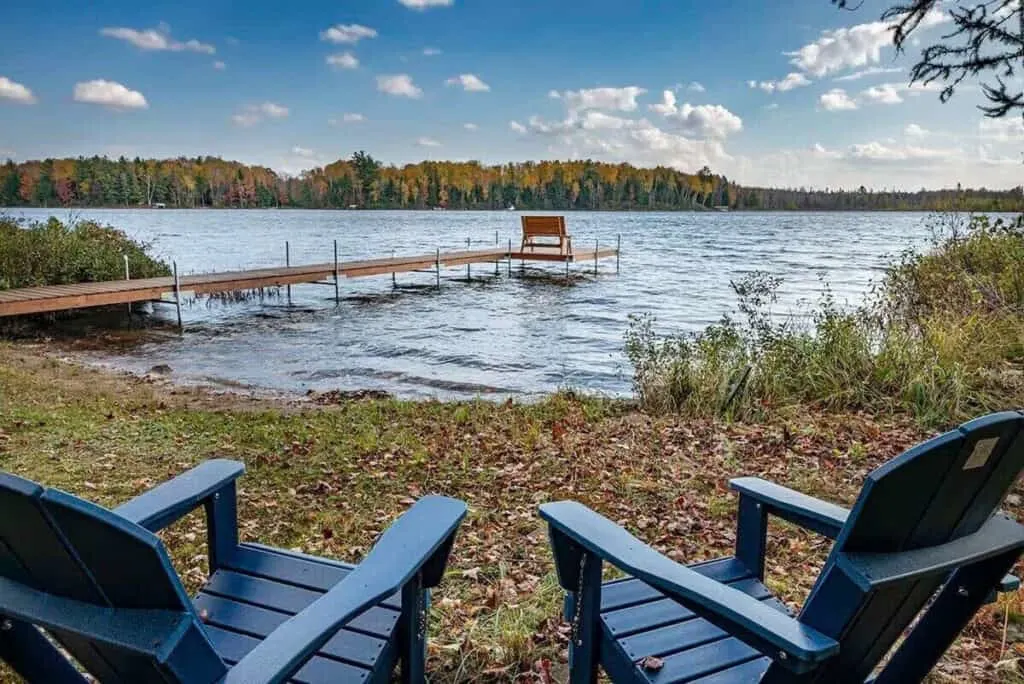 top luxury vacation rentals in wisconsin, Two wooden chairs facing a wooden jetty standing on the shores of a lake surrounded by trees in fall colors under a wide open blue sky with white fluffy clouds