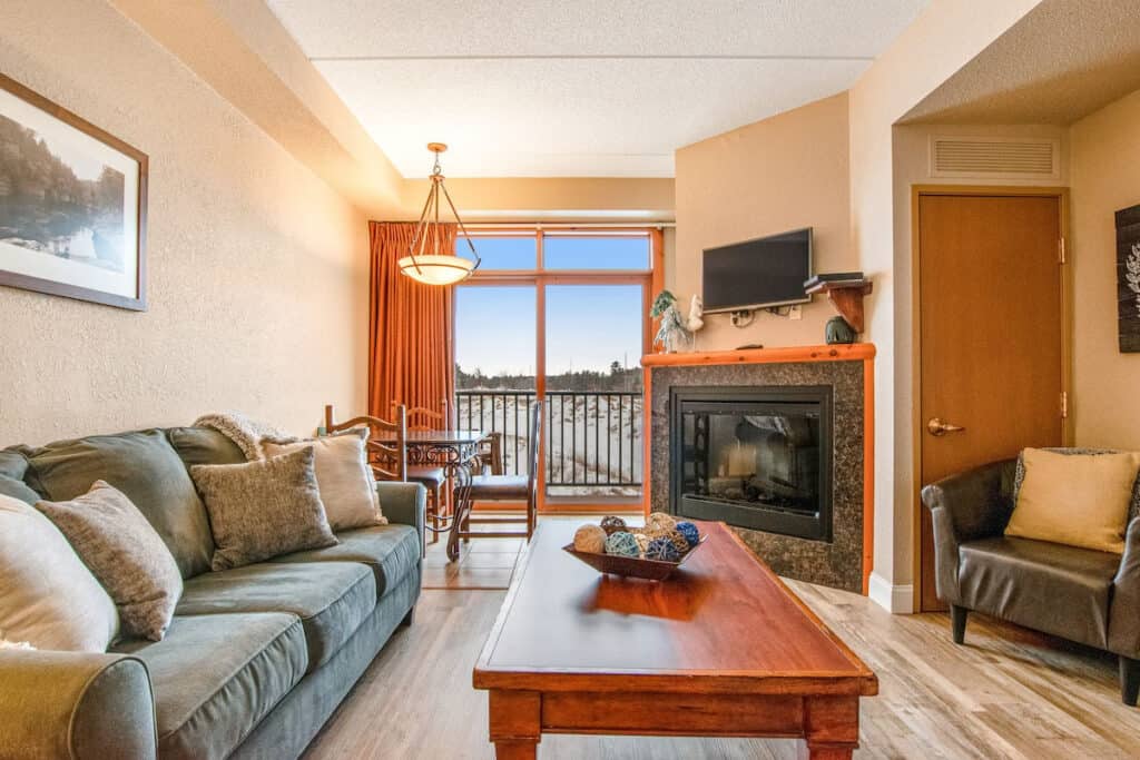 luxury cabins in wisconsin dells, Interior living room with soft comfortable couch next to long low wooden table and wood burning fireplace with armchair to one side and a balcony visible behind