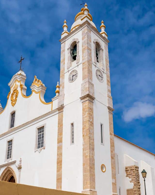 Upward view of a tall stone clock tower attached to a white stone church under a blue sky with wispy white clouds