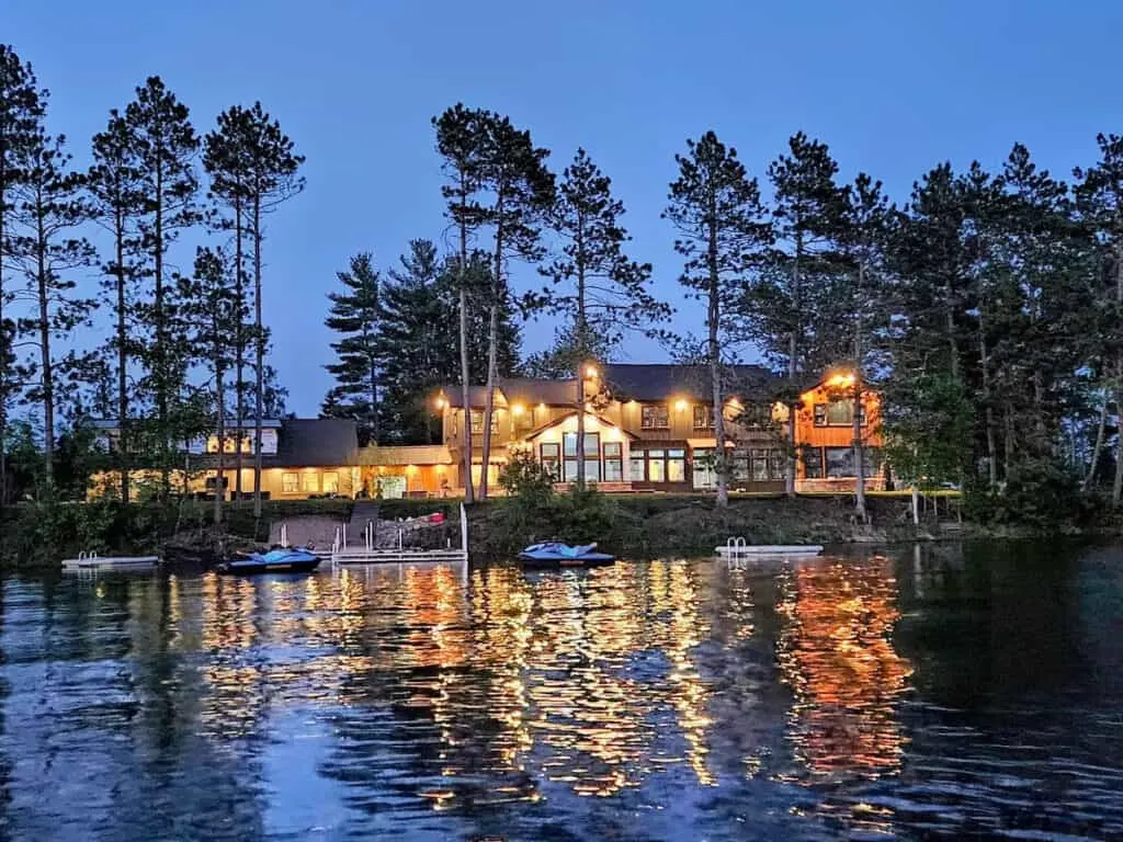 wisconsin luxury cabin rentals, View from lake of large luxury cabin buildings lit by warm outdoor electric lights surrounded by tall trees and jet skis moored by a wooden jetty at dusk