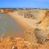 Amazing things to do in Sagres Portugal, View of golden sandy beach with small waves making white surf on the shore all surrounded by small rocky cliffs with buildings visible on the horizon under a clear blue sky