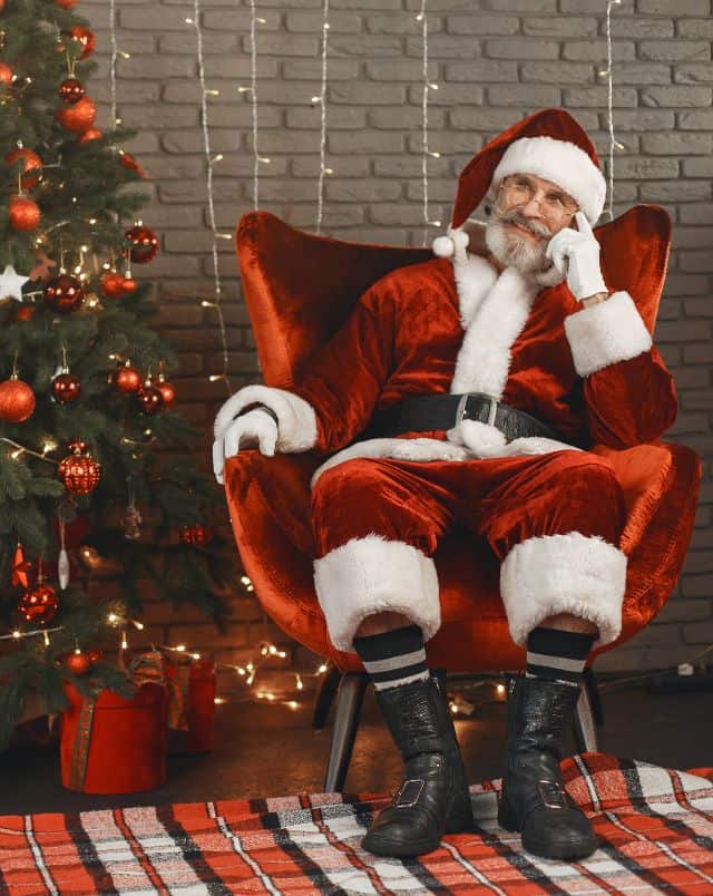 fun things to do in Milwaukee in December with kids, Smiling Santa Claus sitting in red chair next to a Christmas tree bedecked in red baubles and stars with electric lights behind him