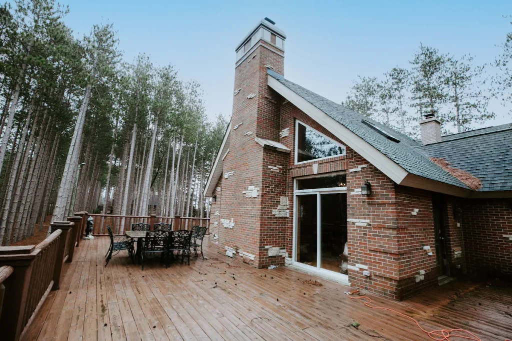 luxury wisconsin cabin rentals, Outdoor wrap around deck with table and chairs surrounding a brick cabin with vaulted ceiling also surrounded by tall green trees under a clear blue sky