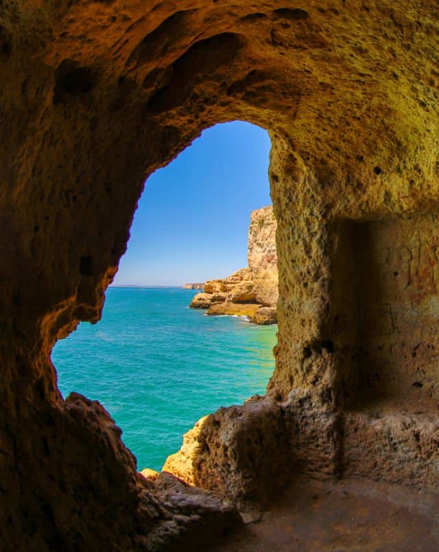 What to do in Carvoeiro, View from inside a small cave through an opening out to sea with rocky cliffs sitting next to the bright turquoise waters of the sea under a clear blue sky