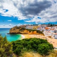Best things to do in Carvoeiro Algarve, View of seagulls flying over a beach with golden sand next to turquoise seawater with rocky cliffs and rows of densely packed residential buildings with white fronts and colorful rooftops all under a blue sky with clouds