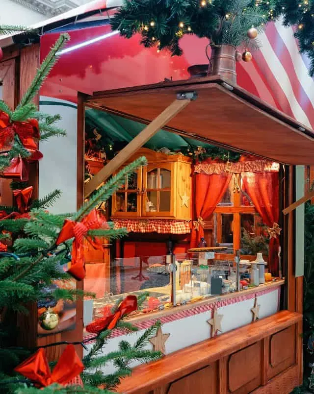 Wisconsin german Christmas market, Christmas market stall with open front revealing glass counter and serving station with wooden cabinet and window with red curtains behind all decorated in Christmas ornaments