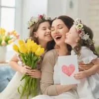 mother kissed by her two daughters wearing flower crowns, one giving flowers and other giving a card with a heart