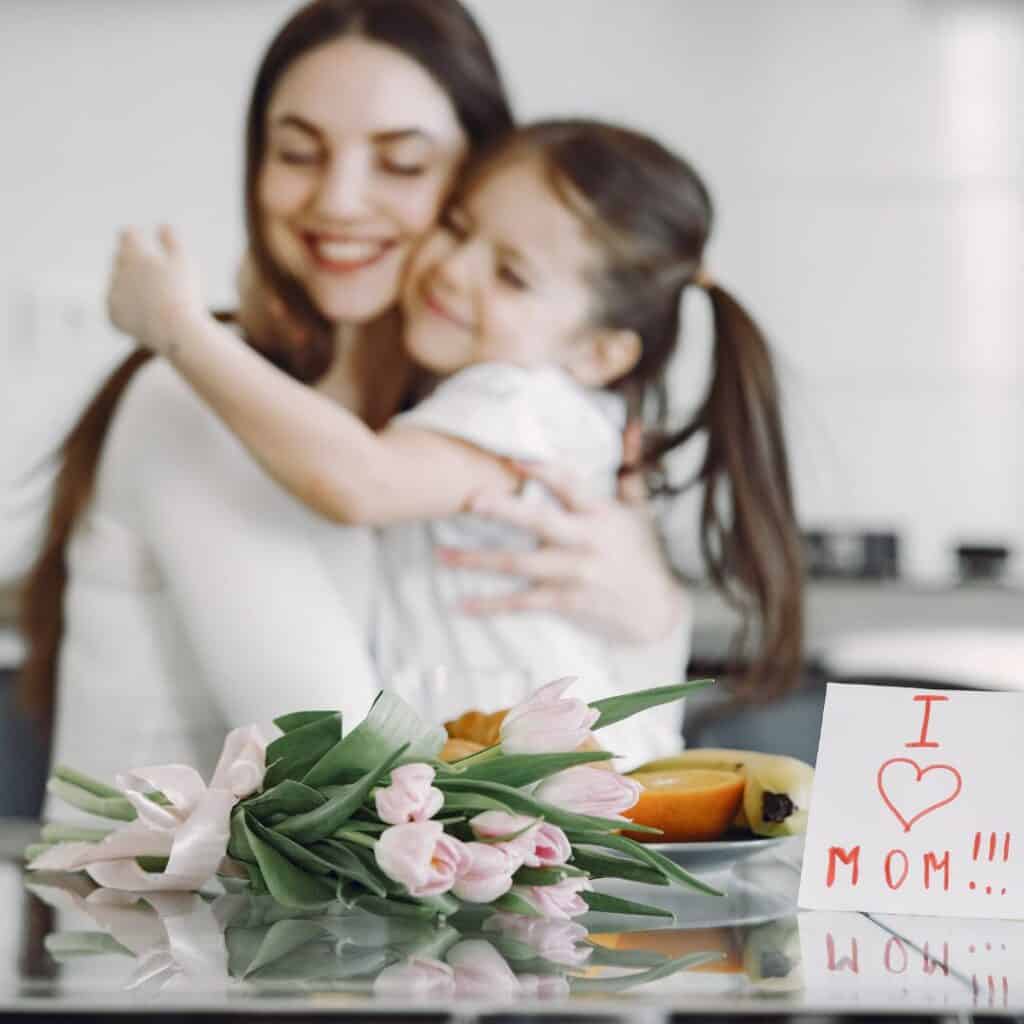 a mom and a daughter hugging tightly with a card saying I love mom and a flower on the foreground