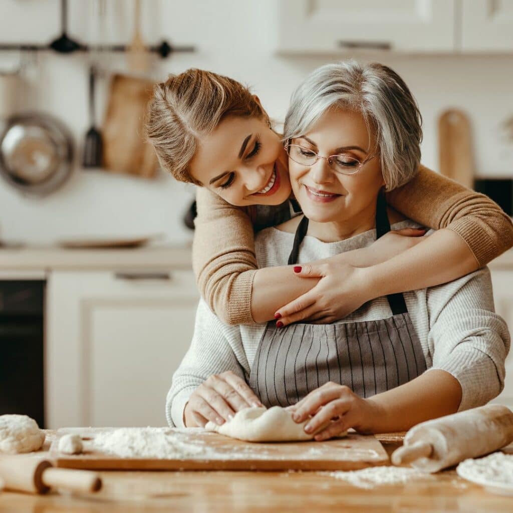 an older mom sitting while making food hugged by her daughter from the back