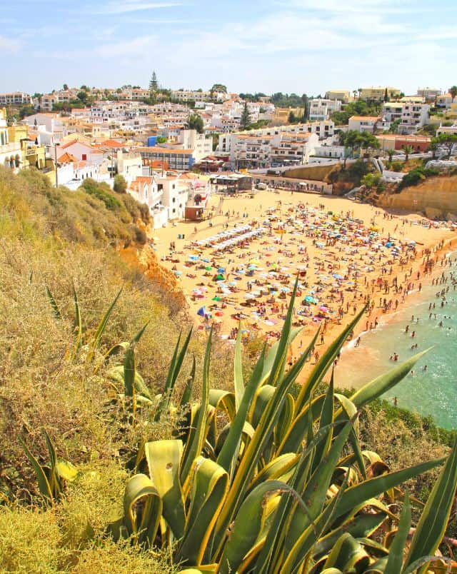 Best beaches in Carvoeiro, View from hillside looking down onto a densely populated beach full of people and beach umbrellas next to a town full of white buildings with colorful rooftops under a bright cloudy sky