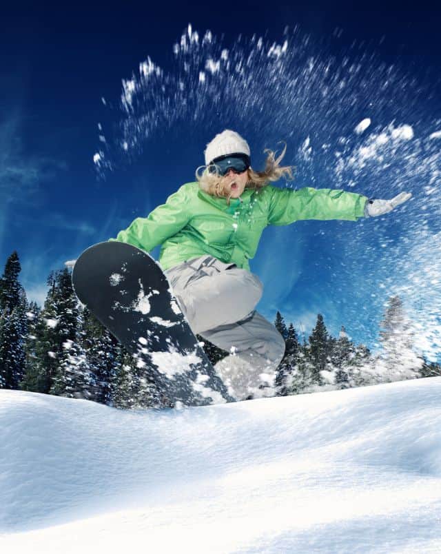 top things to do in Wisconsin Dells in January, Freezeframe of person in green jacket and white hat and goggles riding a snowboard over a hill with white snow spraying around them and a line of green trees behind all under a blue sky