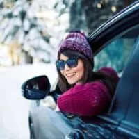 Fun day trips from Milwaukee in winter, Smiling woman wearing sunglasses and purple woolen winter clothes leaning out of the open window of a car with snow-covered trees behind