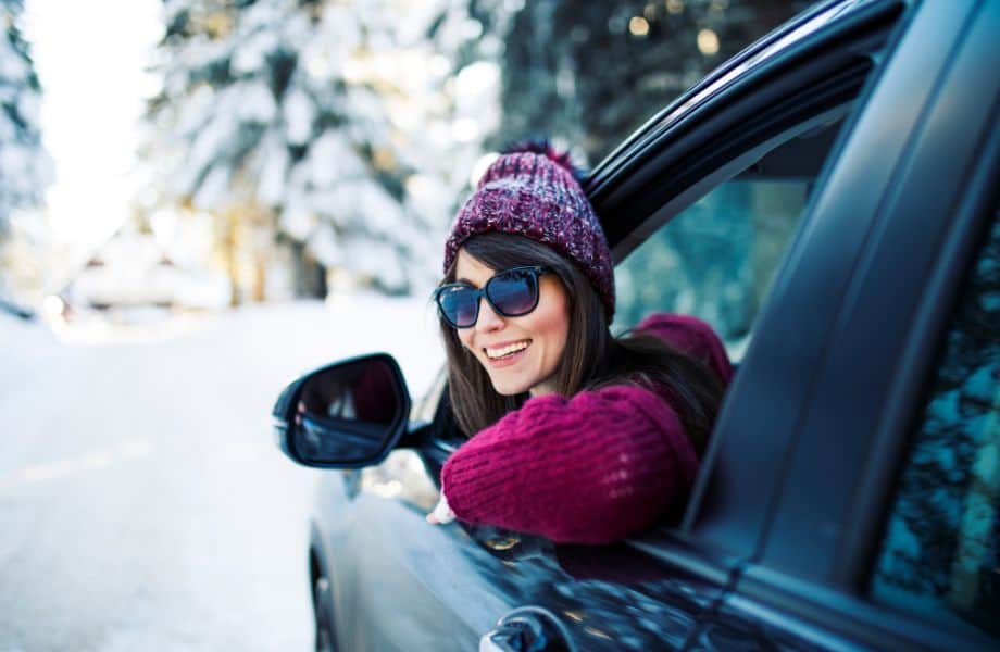 Fun day trips from Milwaukee in winter, Smiling woman wearing sunglasses and purple woolen winter clothes leaning out of the open window of a car with snow-covered trees behind