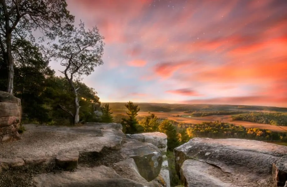 Wisconsin family vacations in nature, Dramatic view of a rocky hillside overlooking a wide valley of fields and trees under a sky full of bright orange and pink clouds at sunset