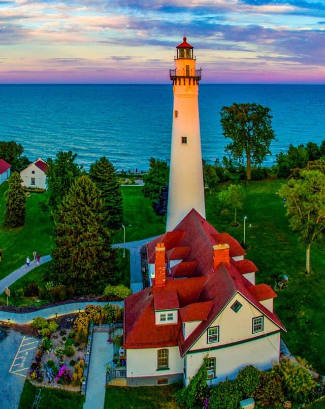 best day trips from Milwaukee in winter, Aerial shot of a lighthouse and its accompanying residential buildings surrounded by green grass and trees with the calm ocean beyond all under a vibrant evening sky with clouds