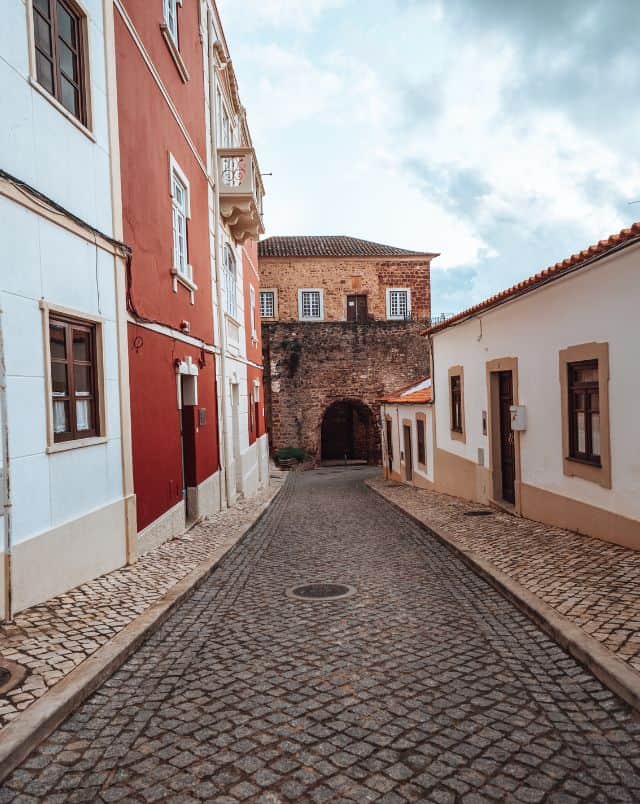 fun activities in Silves, View of an empty cobbled street with low white building on one side and taller red and white apartment buildings on the other with a stone archway at the end of the street