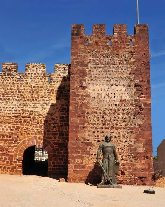 what to do in Silves, Brick tower and wall of Silves Castle with small arched doorway to one side and a bronze statue of a man holding a sword to the other all under a deep blue sky
