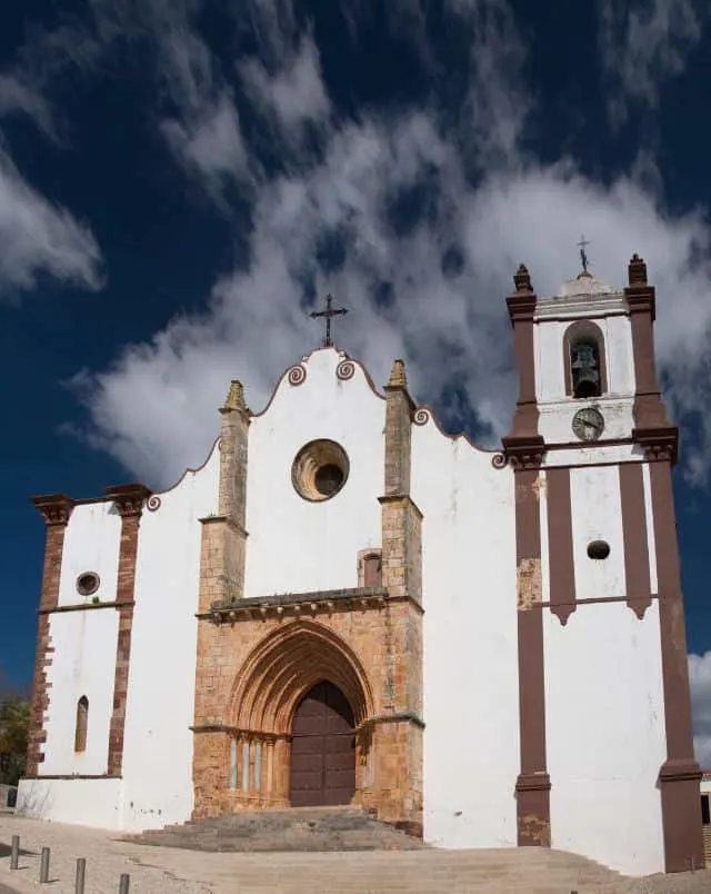 what to see in Silves, View of front of Silves Cathedral with large arched stone doorway surrounded by tall white walls with a bell tower to one side and a crucifix on the top all under a blue sky with white wispy clouds
