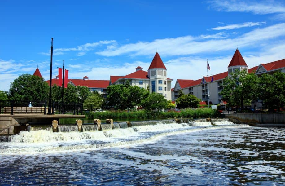 April getaways in Wisconsin, View of buildings with white walls and brown roof tiles with conical towers sitting on the banks of a river running over a small waterfall with green trees lining the shores all under a blue sky with white clouds