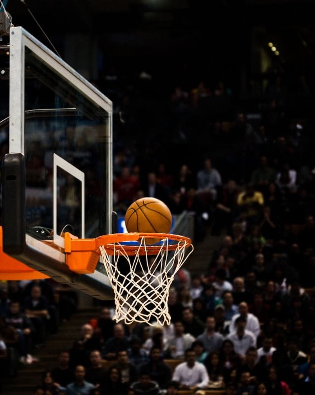 fun things to do in Wisconsin in March, Close up shot of a basketball poised to fall through a basketball hoop in front of a crowd of highly invested sports fans in the background