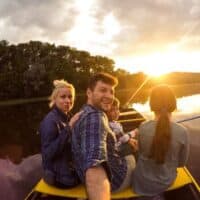 things to do in Wisconsin with kids, Selfie shot of a smiling family of four sitting in a rowing boat and fishing on a clear still lake with a row of trees behind and the bright setting sun on the horizon under a bank of clouds