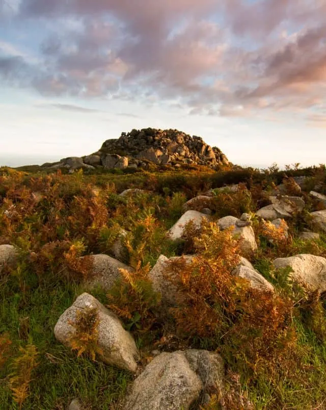 Best Monchique mountains, View of a large mound of earth covered in rocks and boulders seen across a field of green grass peppered with amber bracken and grey stones all under a dramatic dusk sky with clouds