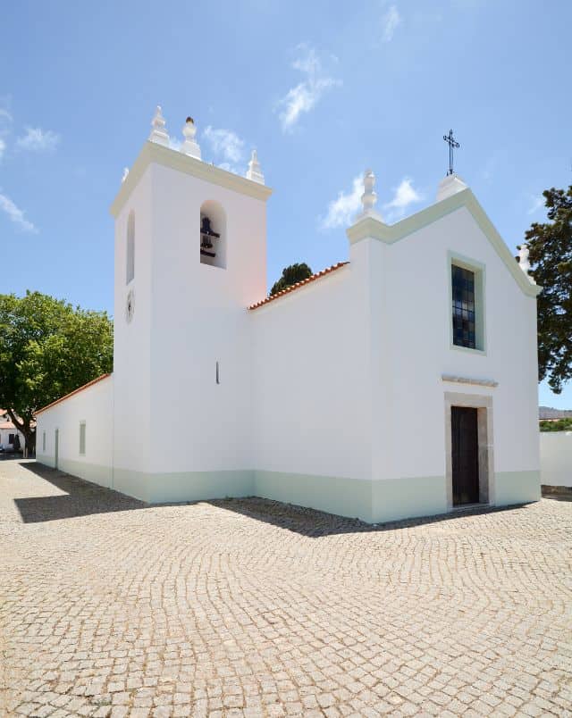 Top Monchique attractions, View of a smooth white church with side bell tower and arched front roof with crucifix on top standing in an open cobbled area with trees behind all under a bright sky with some wispy white clouds