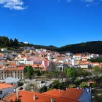 Amazing Things to Do in Monchique, View of a hillside town with white houses with terracotta rooftops built up to a ridge of dark green trees all under a vibrant azure blue sky with some wispy white clouds to one side