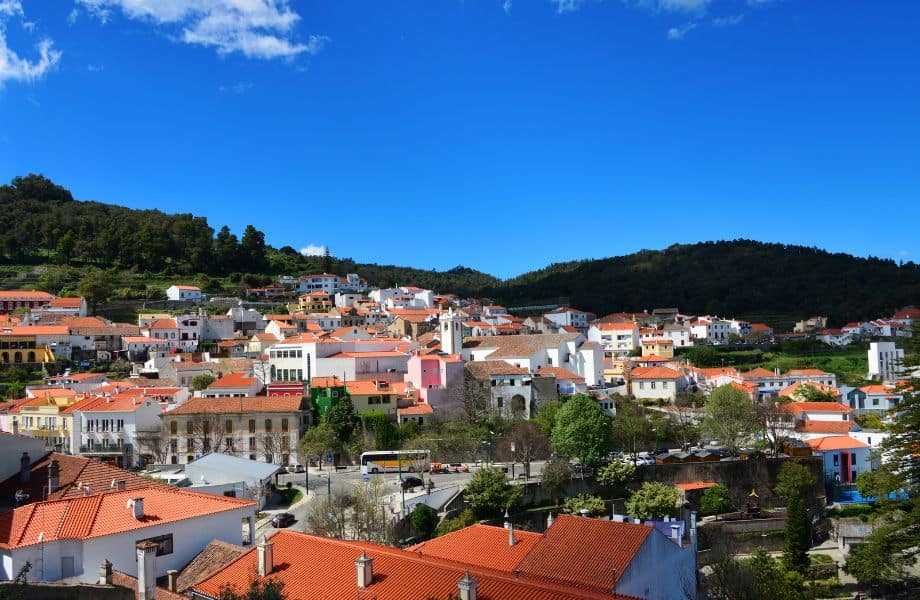 Amazing Things to Do in Monchique, View of a hillside town with white houses with terracotta rooftops built up to a ridge of dark green trees all under a vibrant azure blue sky with some wispy white clouds to one side