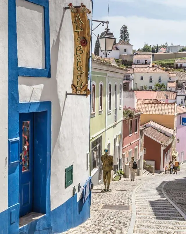 what to see in Monchique, View of people walking down a steep cobbled street with shops and residential buildings to one side decorated in bright soft colors with other buildings lining the side of a hill in the distance all bathed in bright sunshine under a bright cloudy sky