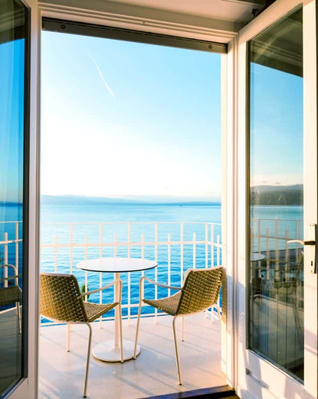 Lake Geneva April activities, View looking out of french windows onto a patio with a small round table and two outdoor wicker chairs with a wide view of a large body of aqua blue water all under a wide open blue sky