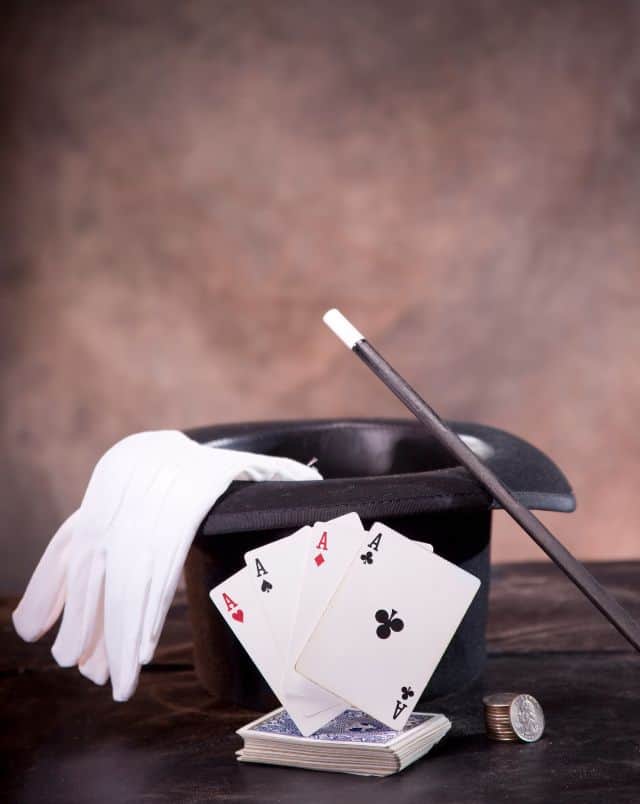 Lake Geneva April events, Close up shot of a magician's top hat with white gloves, a black and white magic wand, a small stack of coins and a deck of cards with the four aces spread out on top