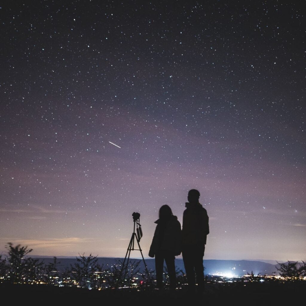 silhouette of two persons stargazing with a telescope on top of a mountain