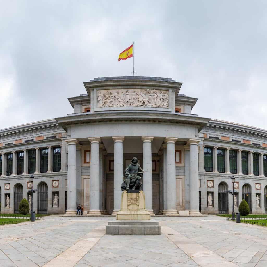 a facade of the Museo Nacional del Prado with greek columns at the entrance and statues and windows, a flag on the roof top and a statue in front
