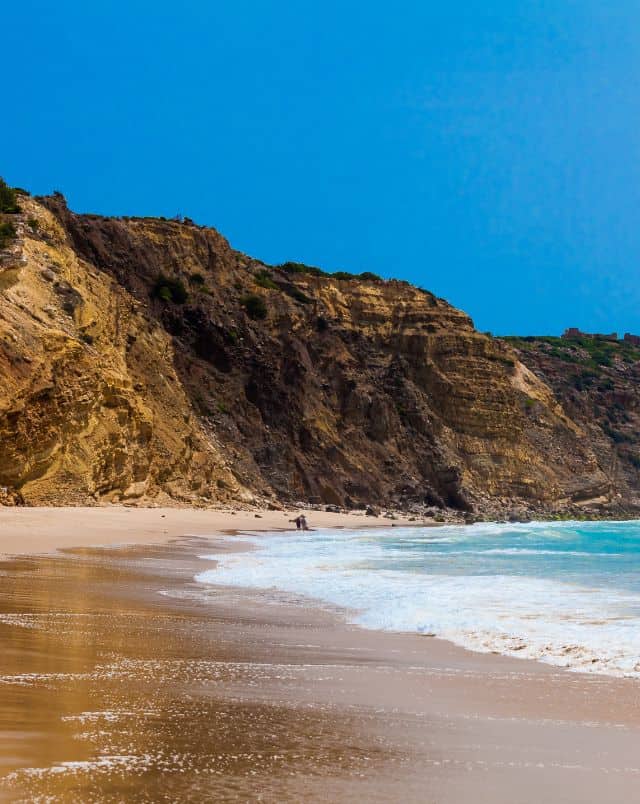 trekking in Algarve, View looking along a sandy beach with blue surf gently crashing on golden sand and tall rocky cliffs behind all under a clear azure blue sky