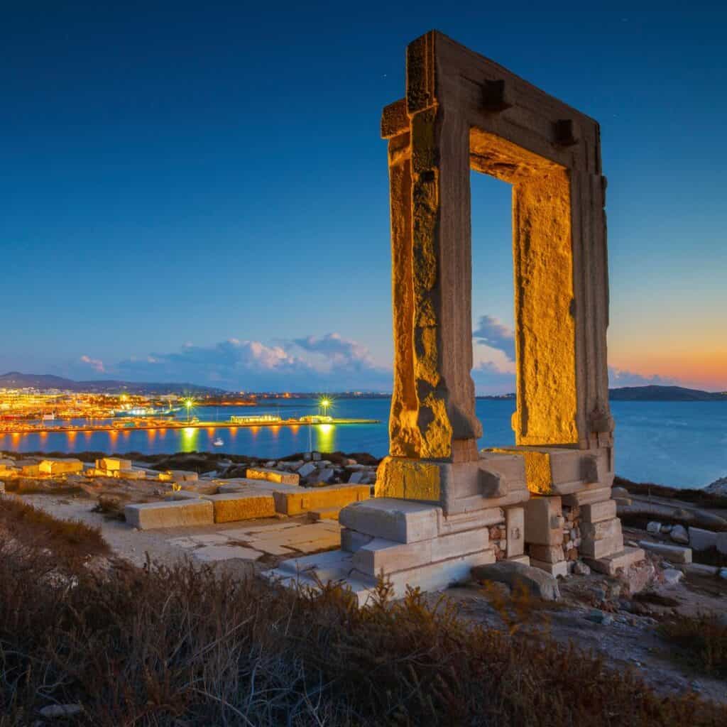 view of Portara and remains of Apollo temple at sunset with the lighted town and the beach at the background