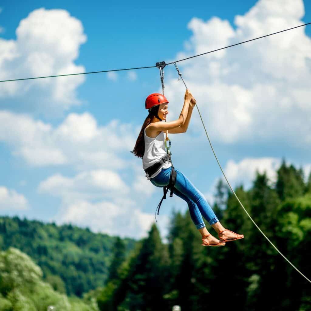 young woman wearing a white top, jeans, sandals, and red helmet riding a zipline in lush forest in lake geneva wisconsin