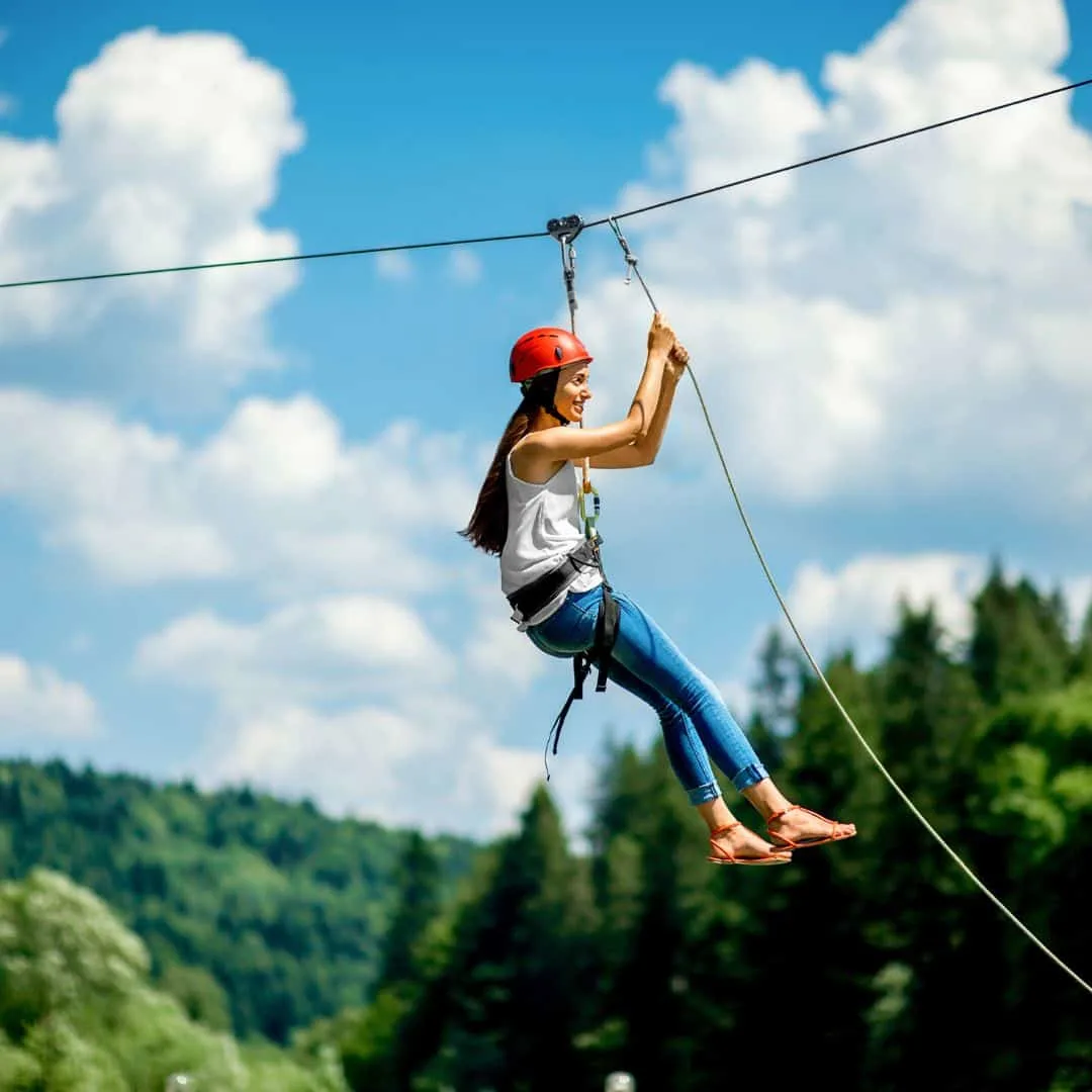 young woman wearing a white top, jeans, sandals, and red helmet riding a zipline in lush forest